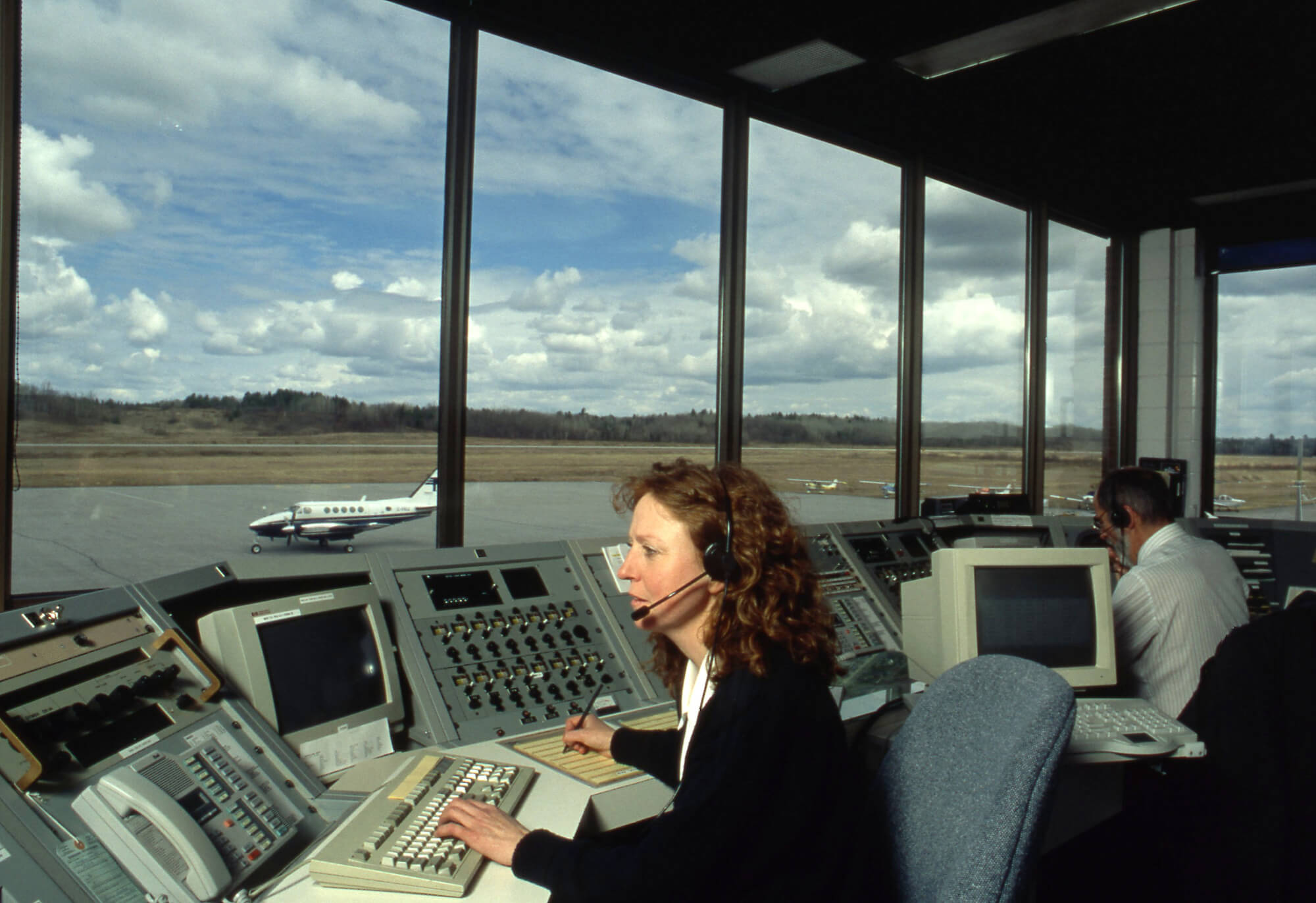 Two people in a tower oversee a small aircraft on the runway. One is speaking into a headset in the background. 