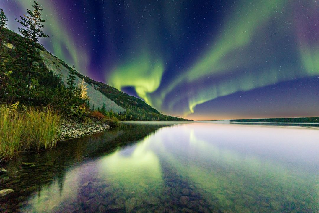 The northern lights glow vibrantly in the sky, reflected in a calm lake beside a forested, rocky shoreline.