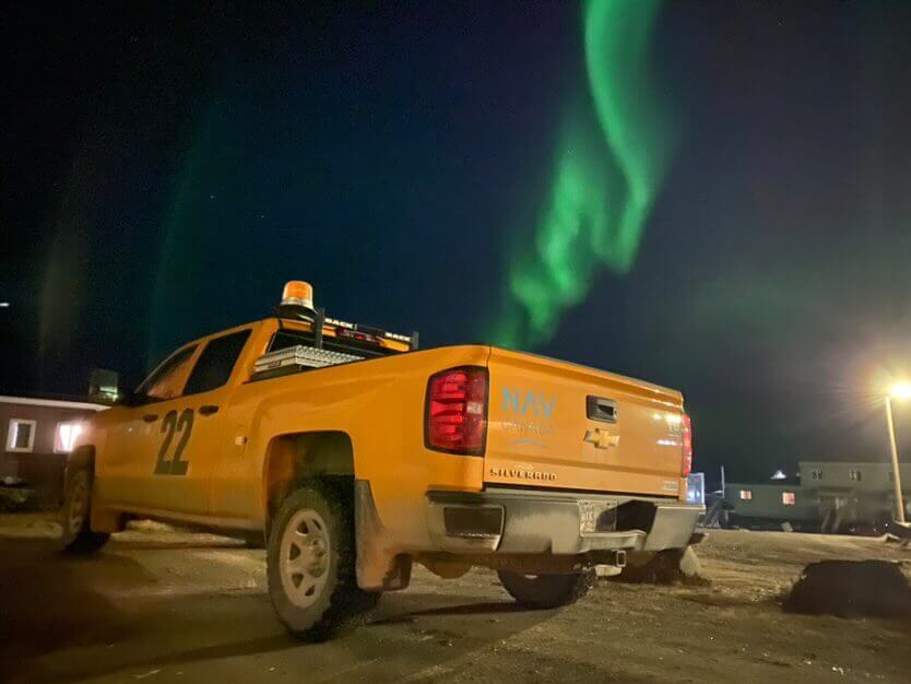 The northern lights shine above a yellow NAV CANADA truck. 